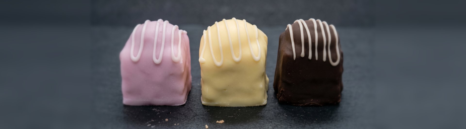 Three fondant fancies, pink, lemon and chocolate, each drizzled with white icing, in a row on dark slate