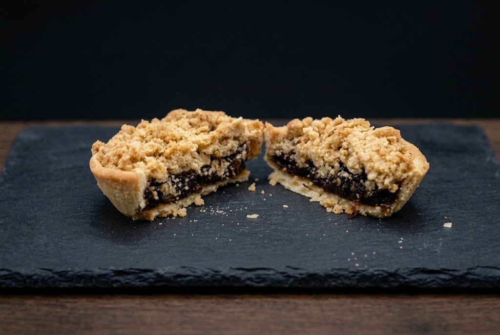 A fruit crumble tart cut in half to show the shortcrust case and dark fruit filling under the crumble
