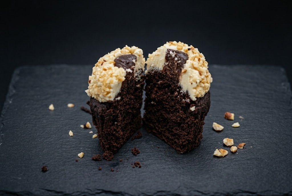 A chocolate cupcake cut in half to show its chocolate centre under a buttercream dome rolled in chopped hazelnuts
