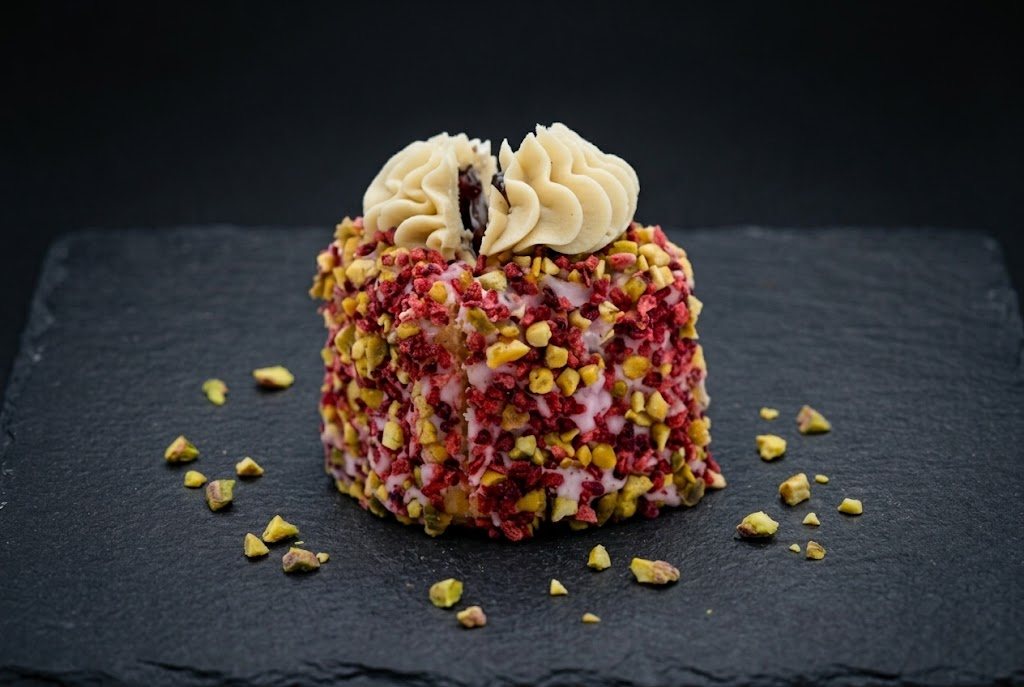 A mini cake coated in chopped pistachio and raspberry pieces, topped with two buttercream swirls and a jam centre