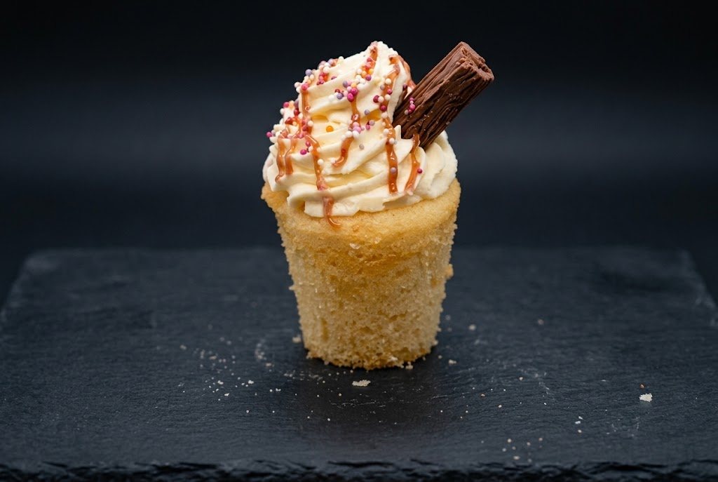 A sundae-style cupcake with a tall cream swirl, sauce drizzle, sprinkles and a chocolate flake, on dark slate