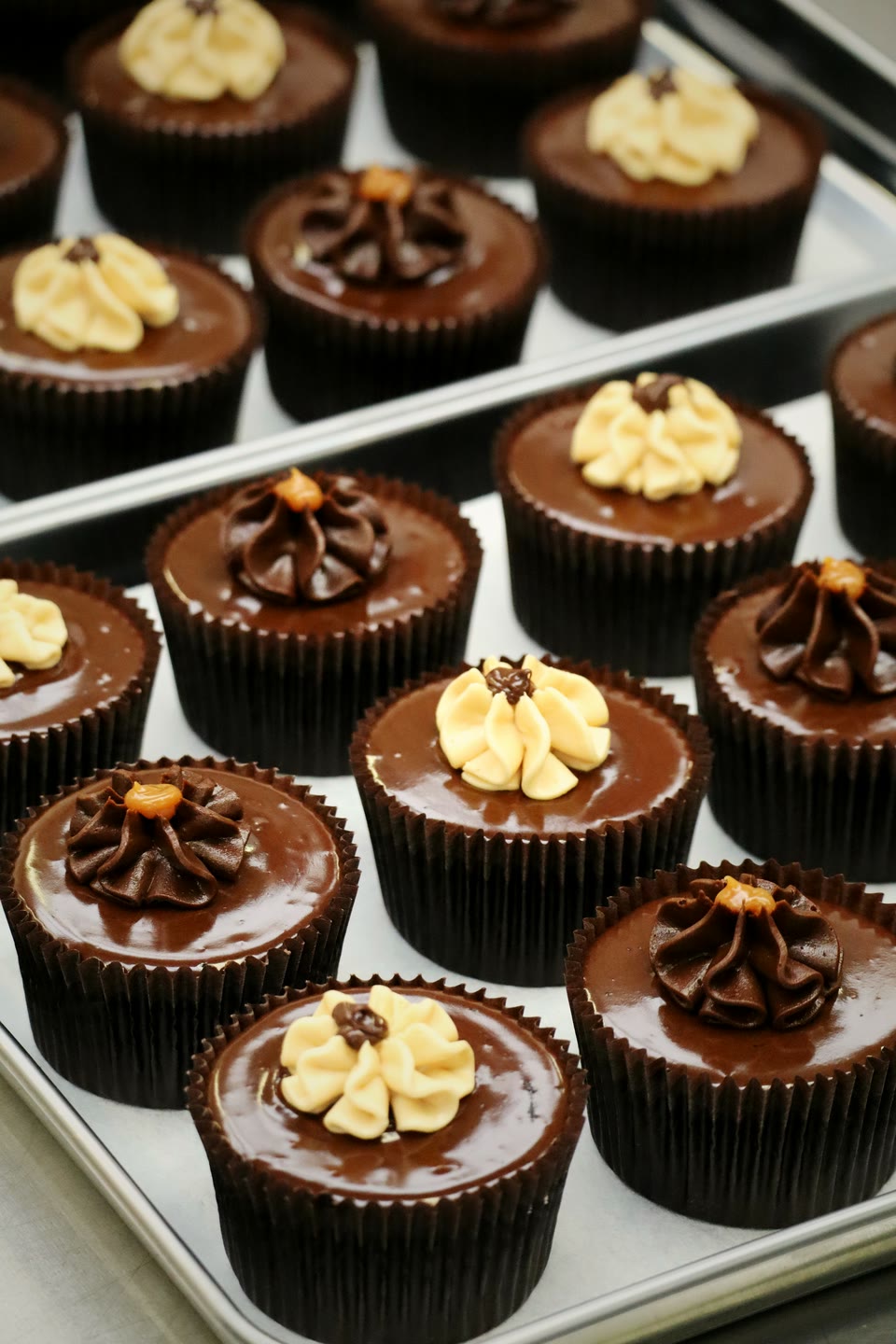 A tray of chocolate cupcakes glazed with ganache, each topped with a hand-piped rosette of caramel or chocolate buttercream