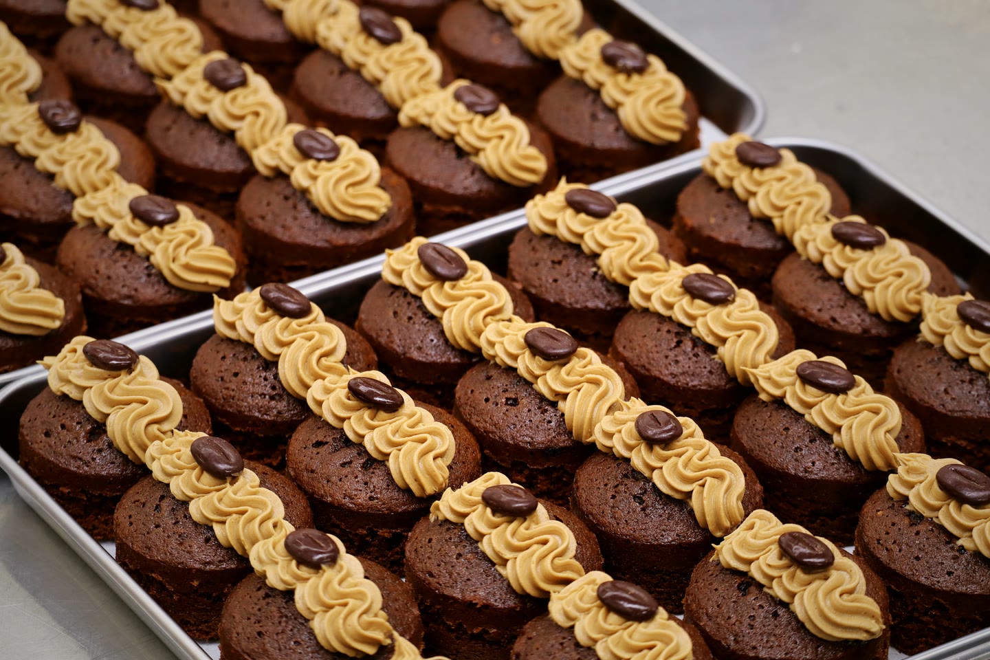 Rows of coffee sponges on a tray, each piped with a ribbon of coffee buttercream and topped with a chocolate coffee bean