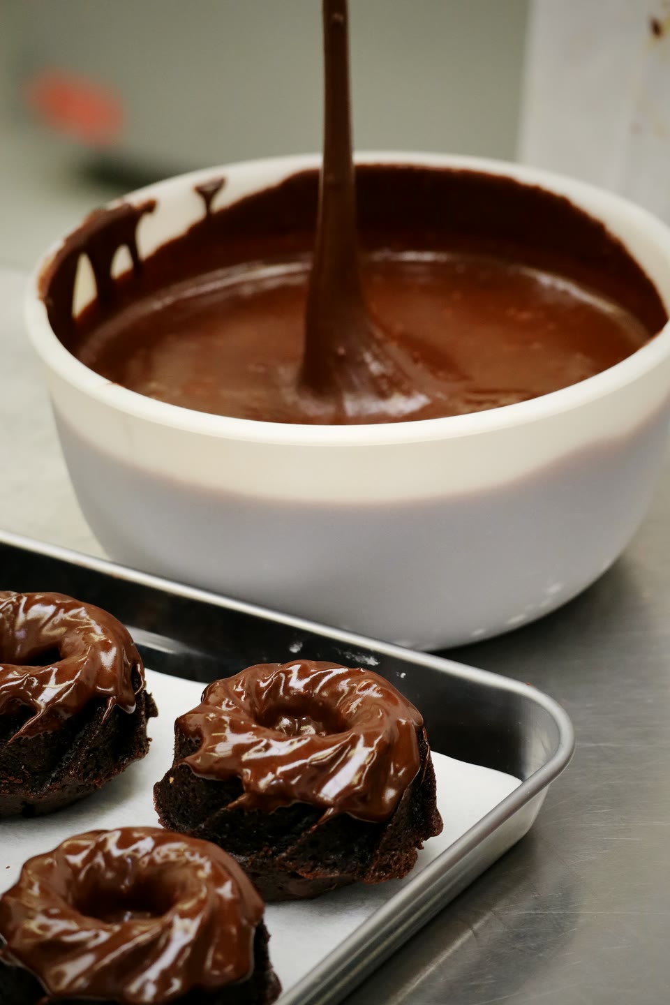 Dark chocolate ganache being poured from a whisk into a bowl, glazed bundt cakes on the tray in front