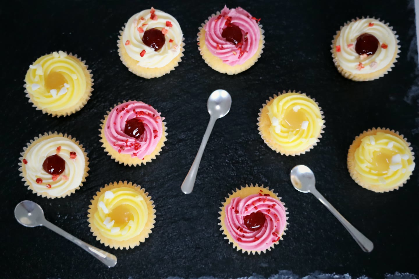 Lemon and raspberry cupcakes arranged on dark slate with silver teaspoons
