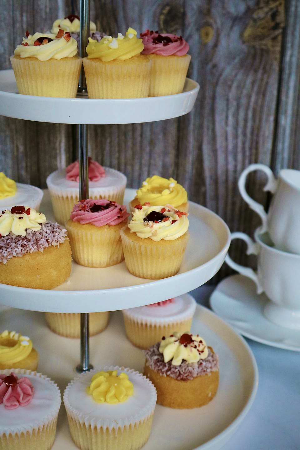 A three-tier cake stand of lemon, raspberry and coconut cupcakes beside a white china tea set
