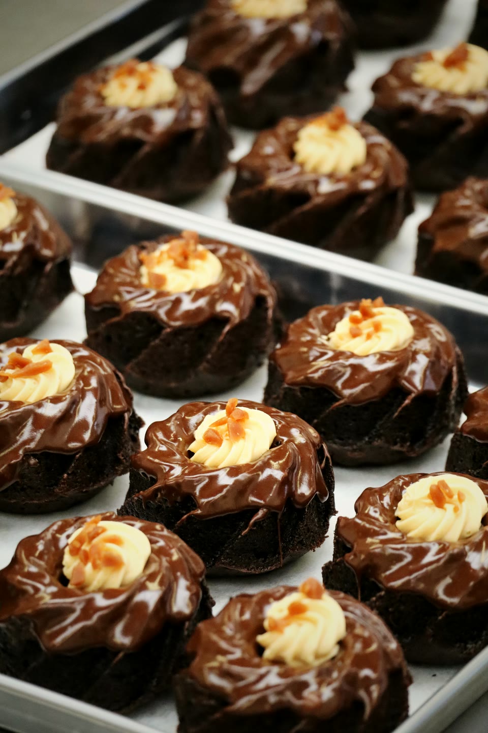 A tray of chocolate bundt cakes glazed with ganache, each finished with a swirl of vanilla cream and a scatter of caramel