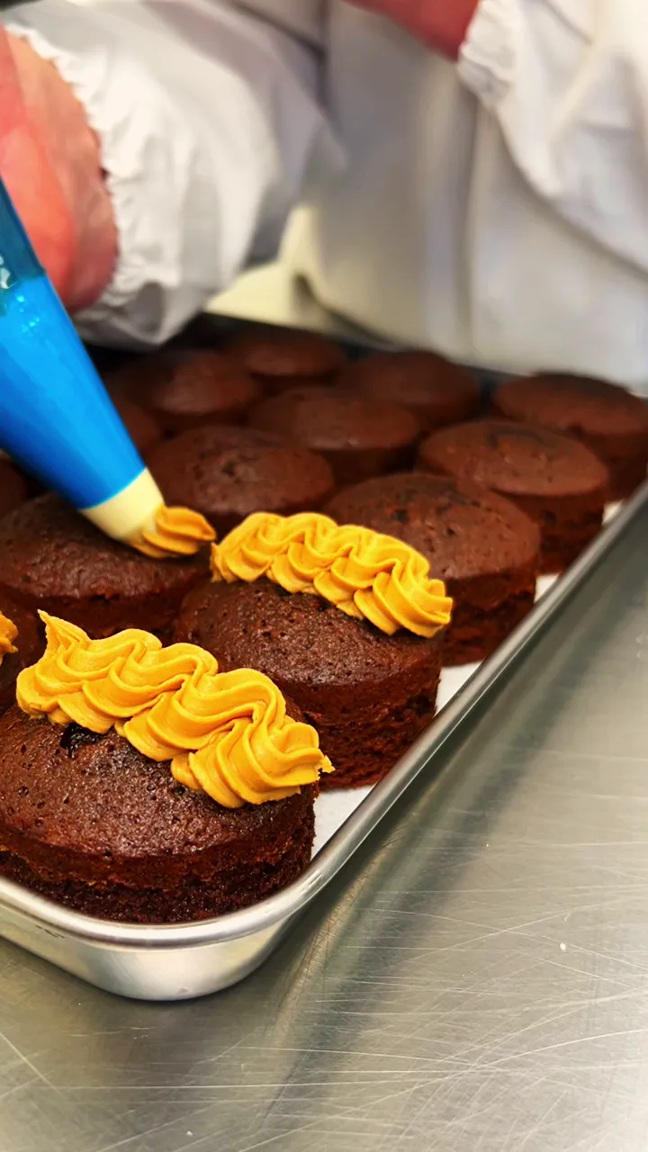 A baker piping caramel buttercream rosettes onto a tray of chocolate cupcakes