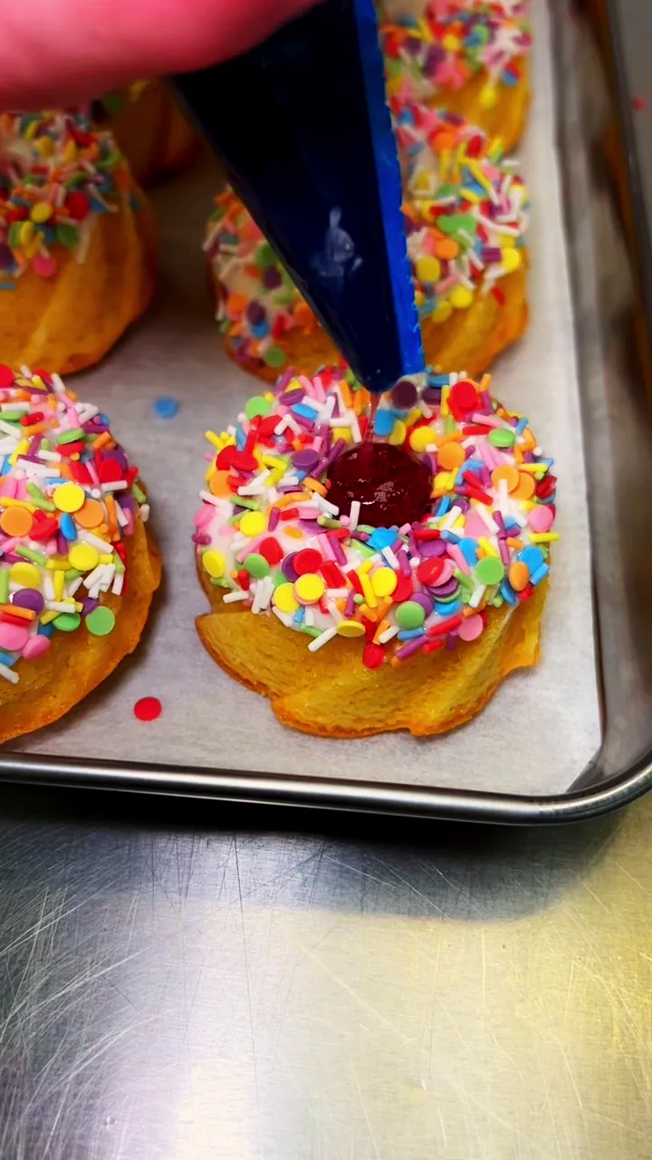 A tray of sponge rings with pink icing and rainbow sprinkles being finished