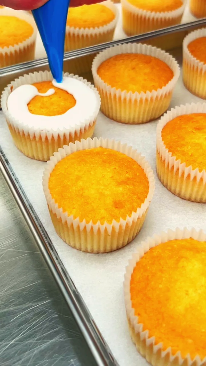 A baker piping white buttercream onto vanilla cupcakes